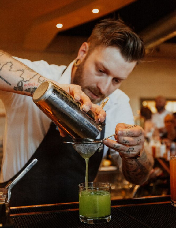 Man pouring a lime green cocktail form the shaker into a glass